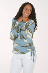 A woman modeling a light blue long-sleeve top with a green and beige leaf pattern, paired with white pants, against a plain white studio background.