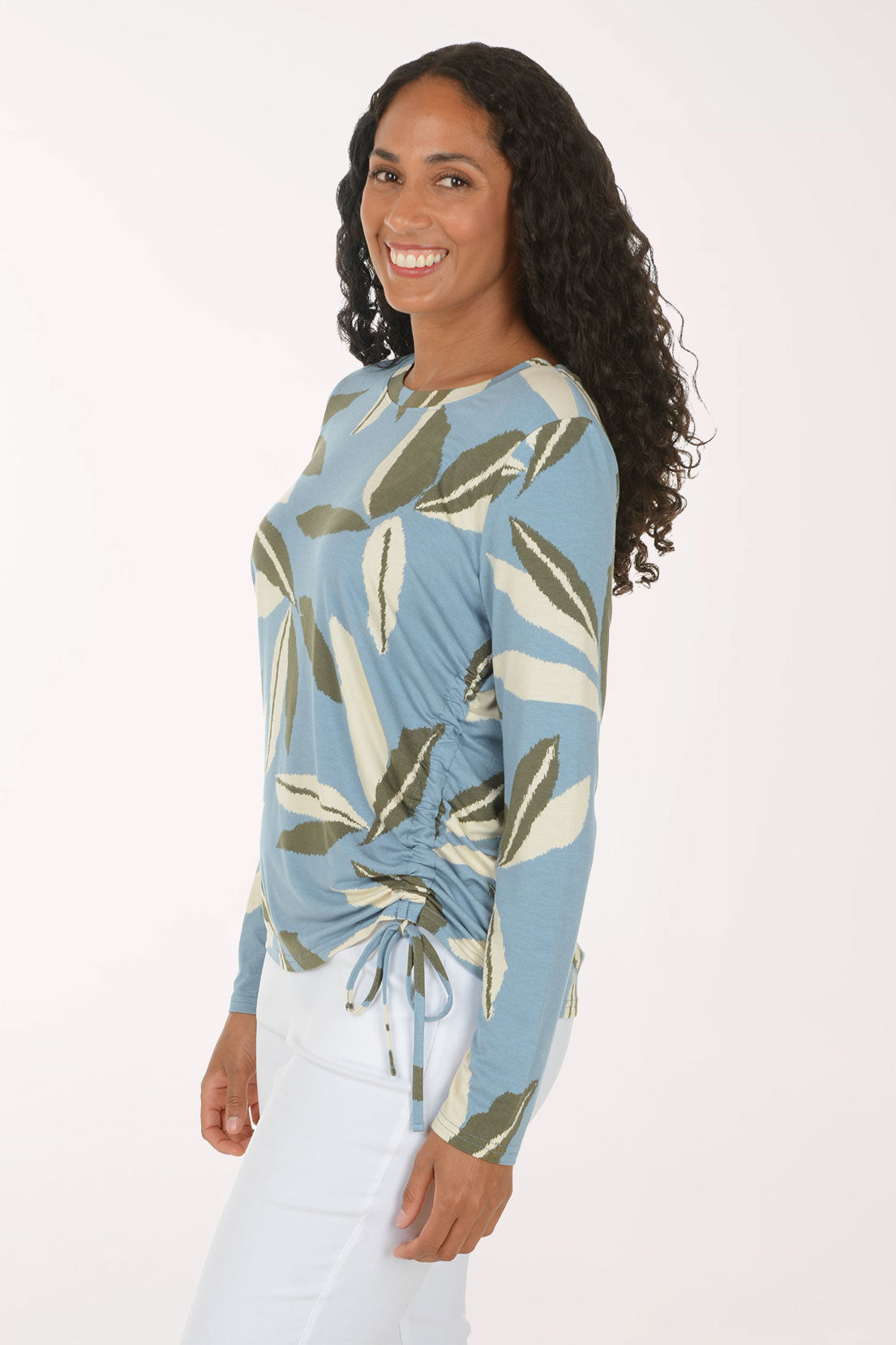A woman modeling a light blue long-sleeve top with a green and beige leaf pattern, paired with white pants, against a plain white studio background.