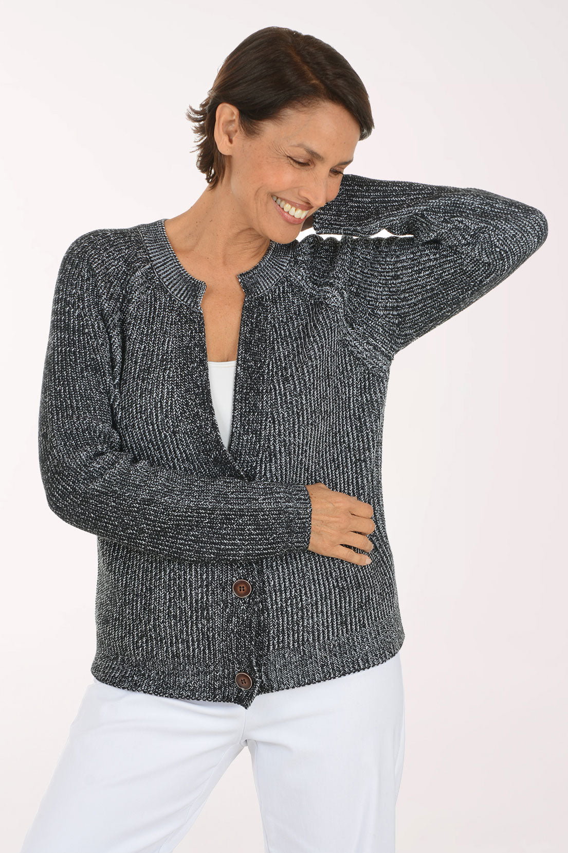 A smiling woman wearing a dark gray and black marled knit cardigan with brown buttons over a white top and white pants, photographed against a plain light background.