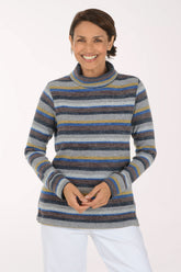 A smiling woman wearing a long-sleeve striped turtleneck sweater with horizontal bands of gray, blue, navy, and mustard yellow, paired with white pants, against a plain white studio background.