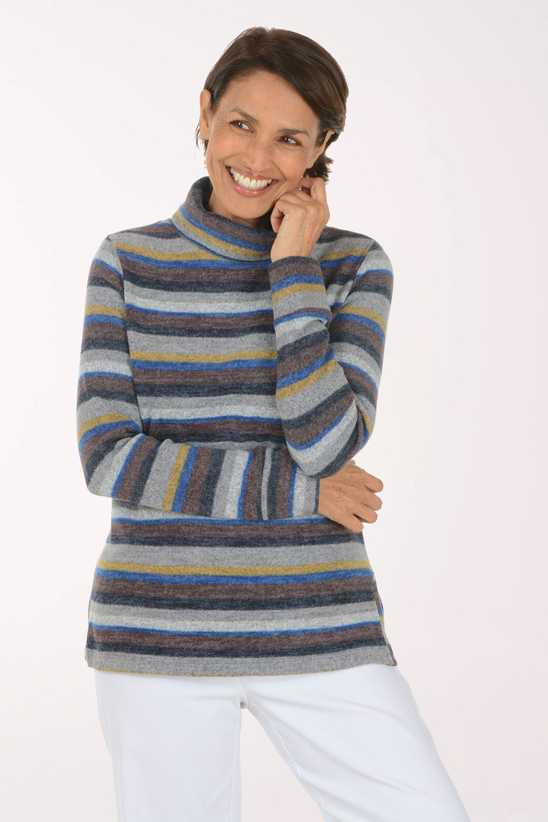 A smiling woman wearing a long-sleeve striped turtleneck sweater with horizontal bands of gray, blue, navy, and mustard yellow, paired with white pants, against a plain white studio background.