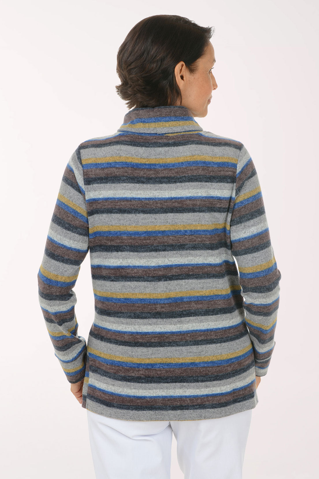 A smiling woman wearing a long-sleeve striped turtleneck sweater with horizontal bands of gray, blue, navy, and mustard yellow, paired with white pants, against a plain white studio background.
