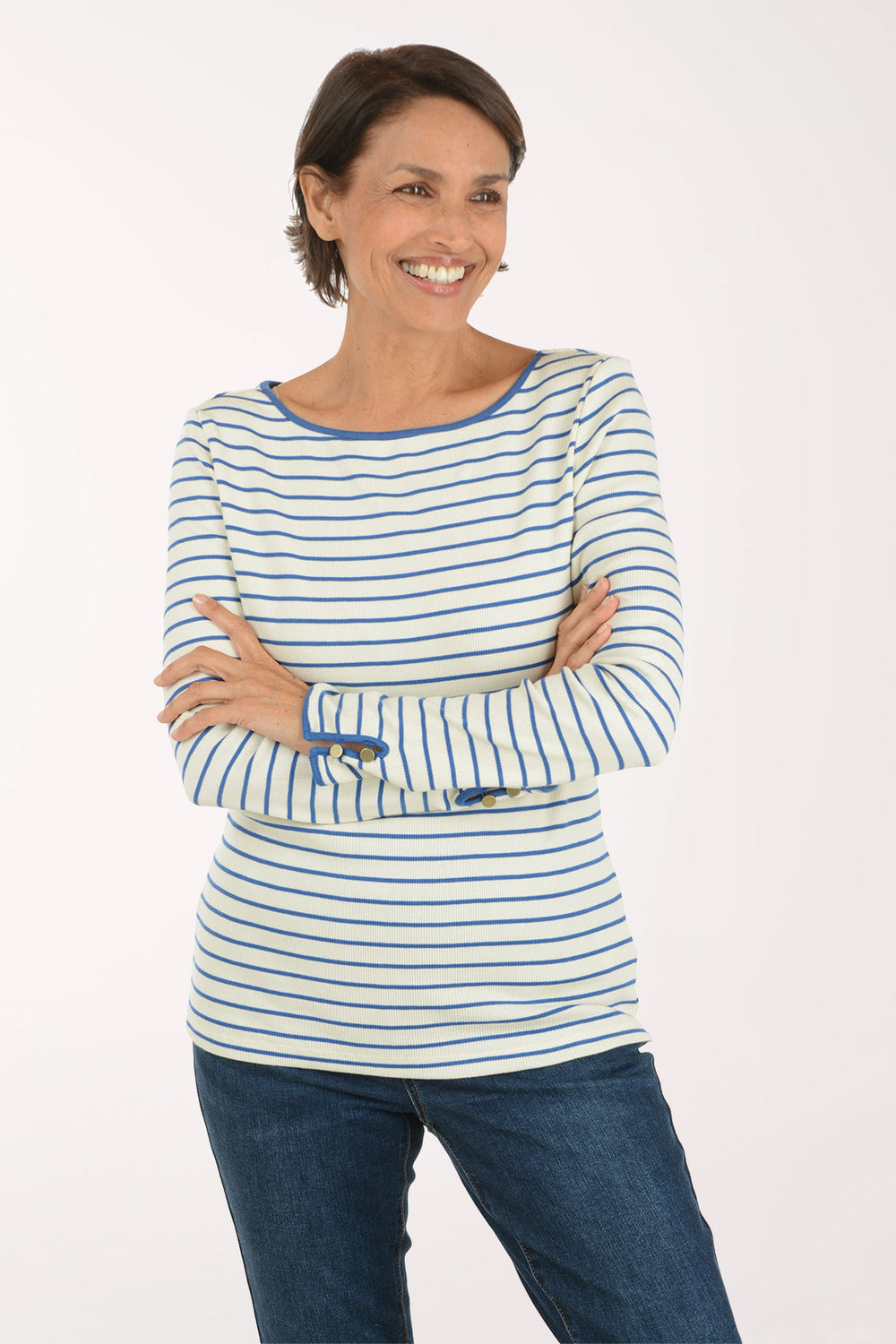 A smiling woman with short brown hair stands with arms crossed, wearing a cream long-sleeve top with thin blue horizontal stripes and blue jeans, against a plain white studio background.