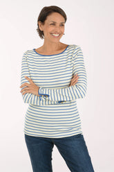 A smiling woman with short brown hair stands with arms crossed, wearing a cream long-sleeve top with thin blue horizontal stripes and blue jeans, against a plain white studio background.