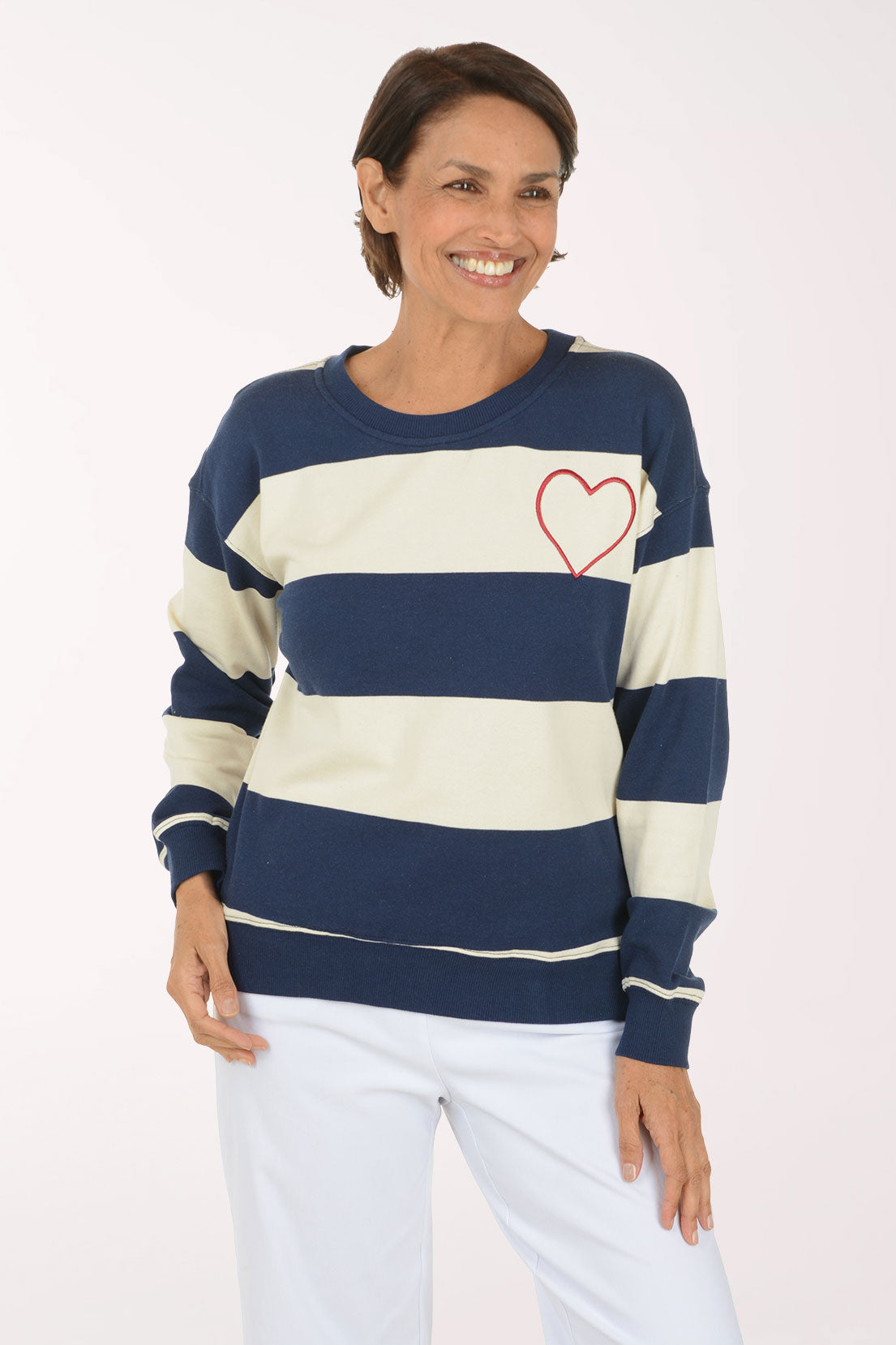 A smiling woman wearing a navy blue and cream striped crewneck sweatshirt with a red heart outline on the chest, paired with white pants, against a plain light background.