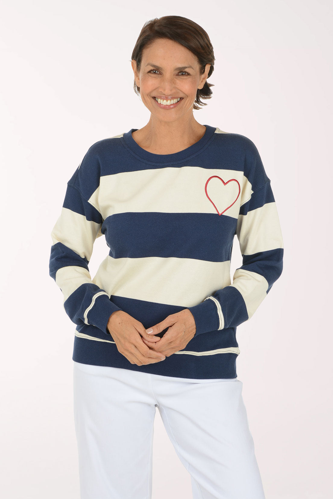 A smiling woman wearing a navy blue and cream striped crewneck sweatshirt with a red heart outline on the chest, paired with white pants, against a plain light background.