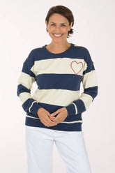 A smiling woman wearing a navy blue and cream striped crewneck sweatshirt with a red heart outline on the chest, paired with white pants, against a plain light background.