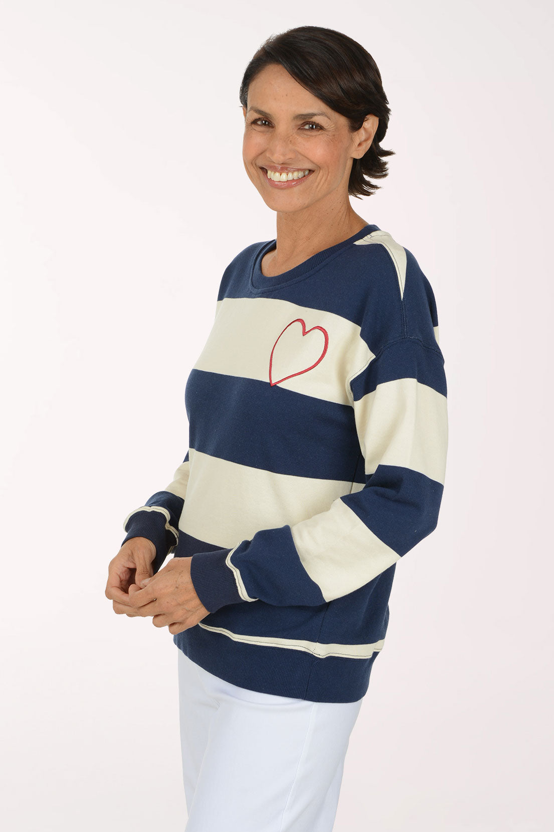 A smiling woman wearing a navy blue and cream striped crewneck sweatshirt with a red heart outline on the chest, paired with white pants, against a plain light background.