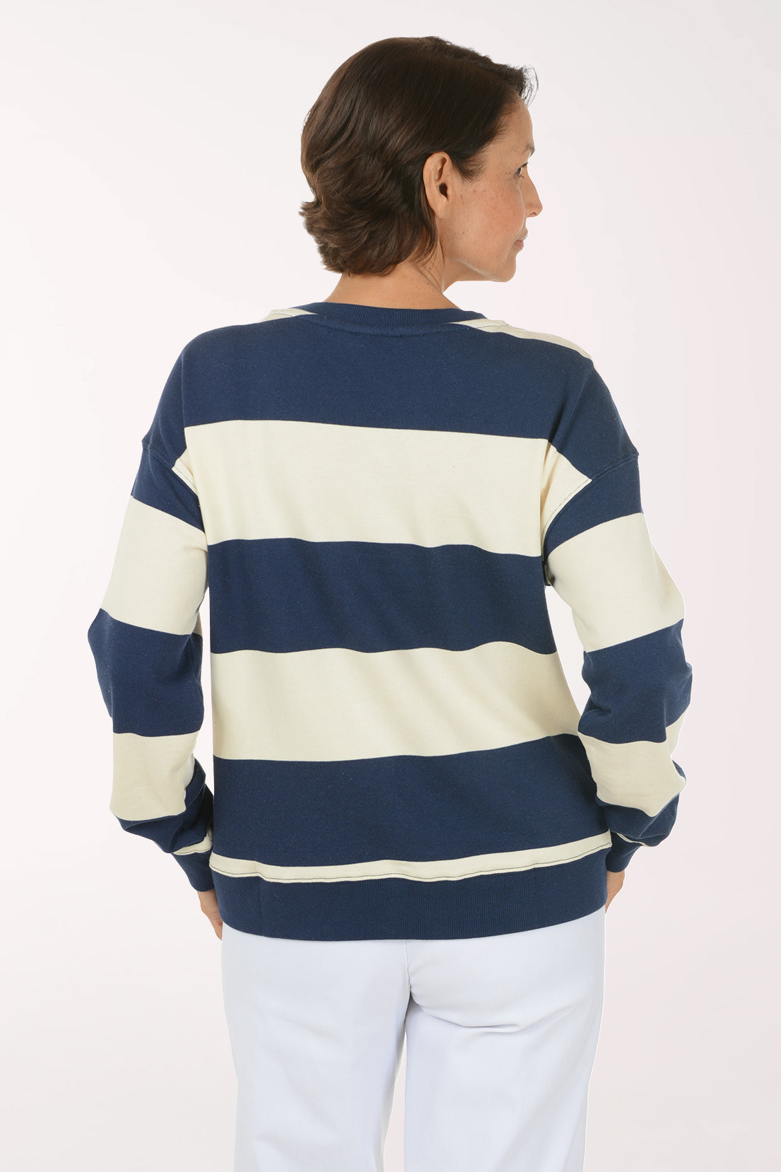 A smiling woman wearing a navy blue and cream striped crewneck sweatshirt with a red heart outline on the chest, paired with white pants, against a plain light background.