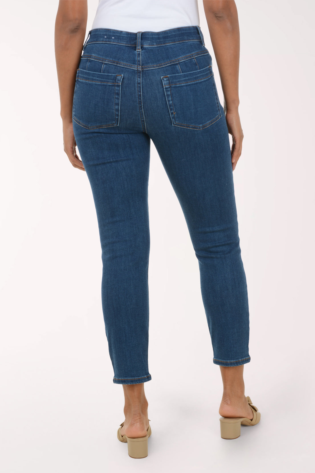 A person shown from the back wearing dark blue skinny jeans with a white top and beige heeled sandals, photographed against a plain light background.