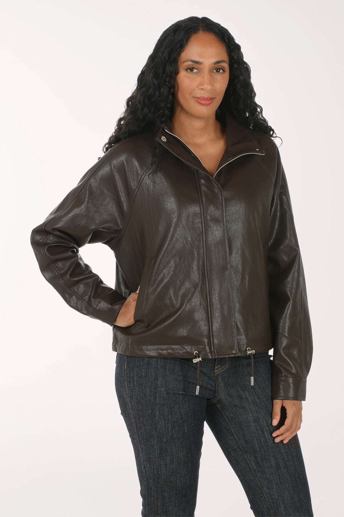 A woman modeling a dark brown faux-leather moto jacket with silver zipper and snap details, worn over a white tank top and dark blue jeans, against a plain white studio background.