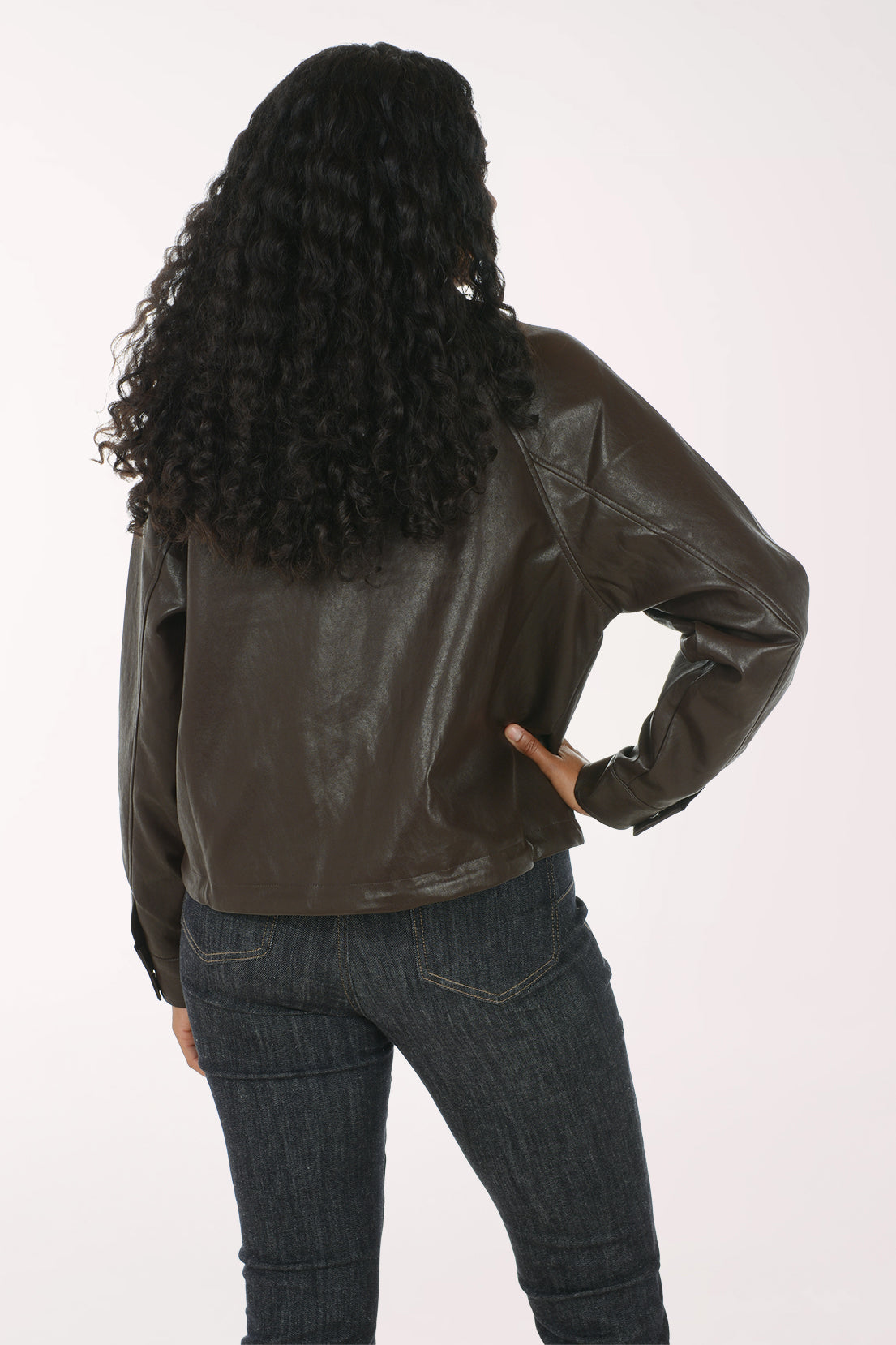A woman modeling a dark brown faux-leather moto jacket with silver zipper and snap details, worn over a white tank top and dark blue jeans, against a plain white studio background.