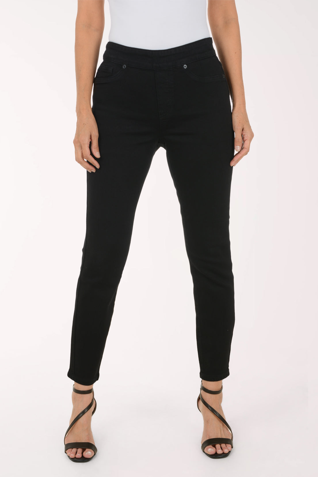 A model wearing black slim-fit ankle-length jeans with a high waist, paired with a white top and black strappy heels, shown against a plain white studio background.