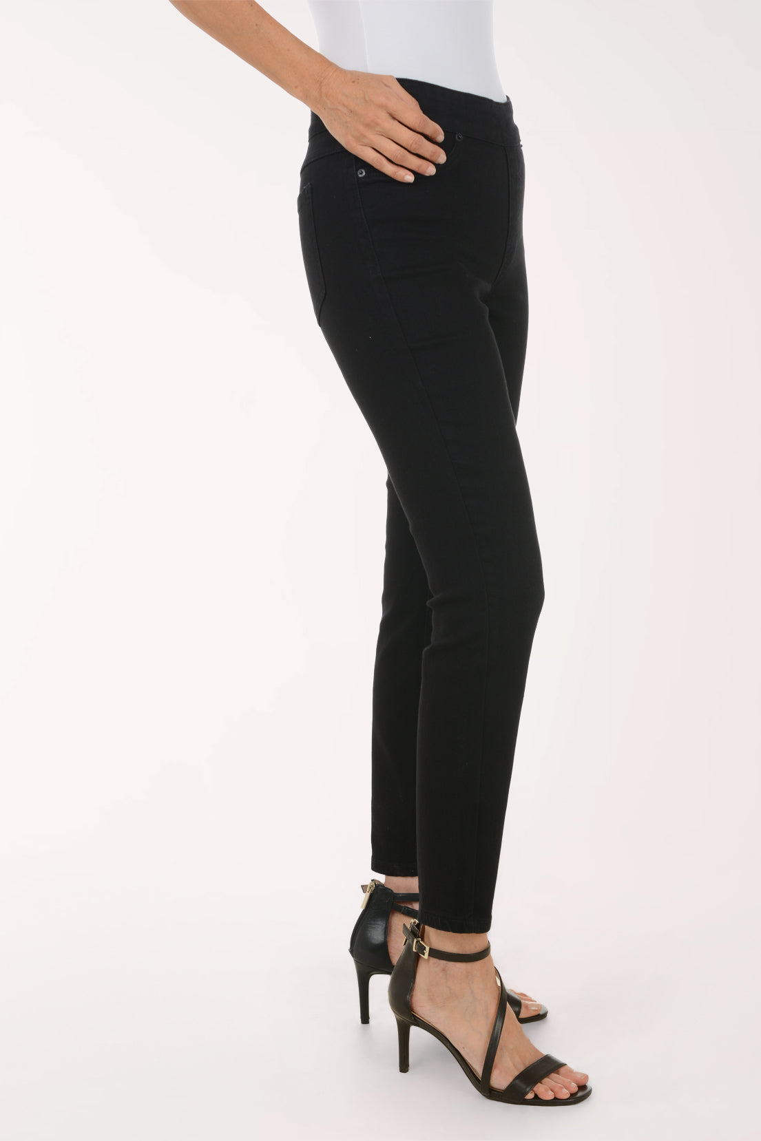 A model wearing black slim-fit ankle-length jeans with a high waist, paired with a white top and black strappy heels, shown against a plain white studio background.