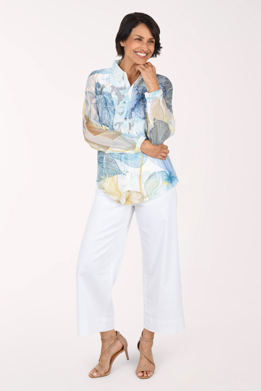 Full-length front view of a woman in white stretch denim wide leg crop pants worn with a long sleeve blue, cream and gold abstract print button-front shirt and nude heeled sandals.