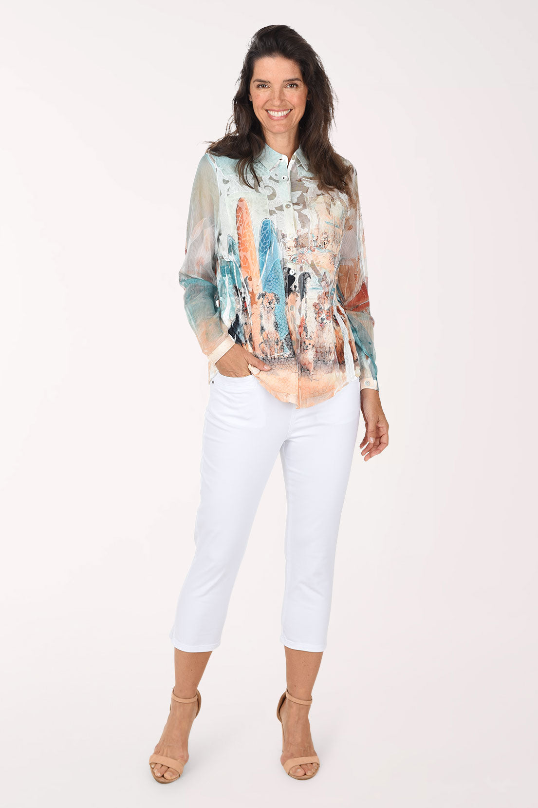 Full-length front view of a woman in white stretch denim capri pants worn with a long sleeve watercolor print blouse in blue, peach and cream and nude heeled sandals.