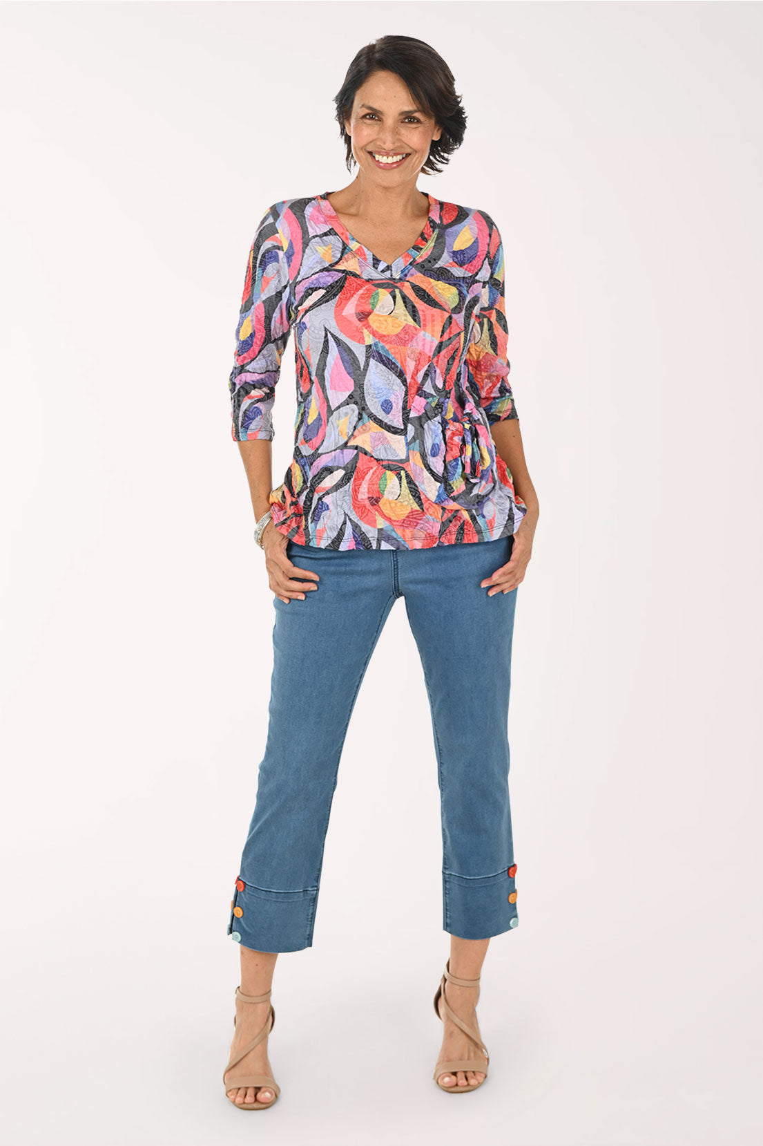 A woman modeling a colorful abstract print long-sleeve top paired with blue cropped jeans and beige heeled sandals, photographed against a plain white studio background.