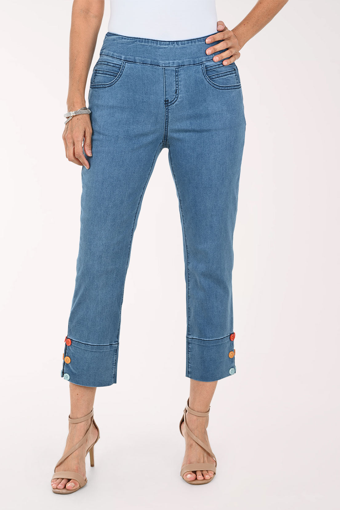 A model wearing blue cropped denim jeans with rolled cuffs featuring colorful button details, paired with beige high-heeled sandals against a plain white studio background.