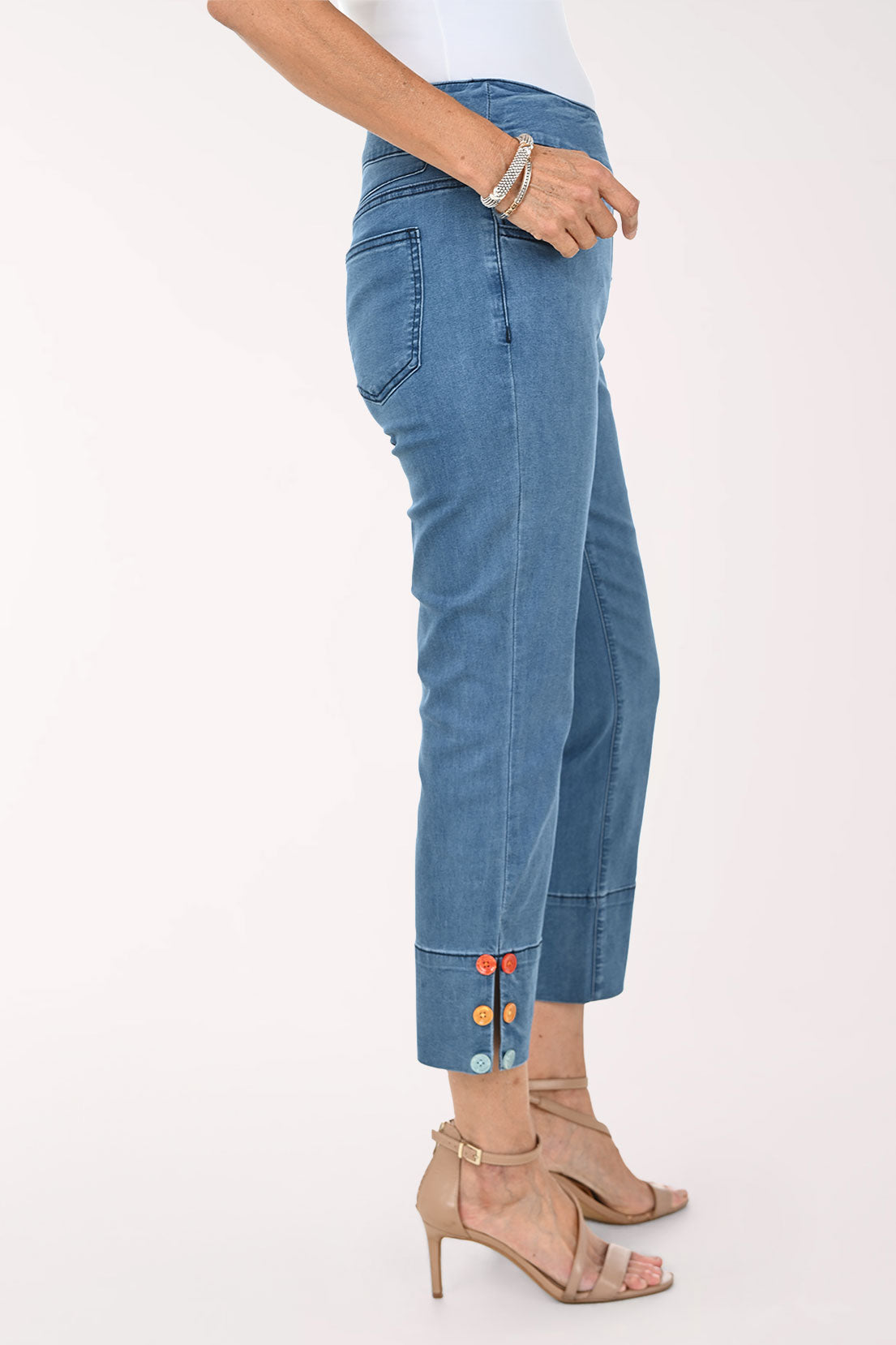 A model wearing blue cropped denim jeans with rolled cuffs featuring colorful button details, paired with beige high-heeled sandals against a plain white studio background.