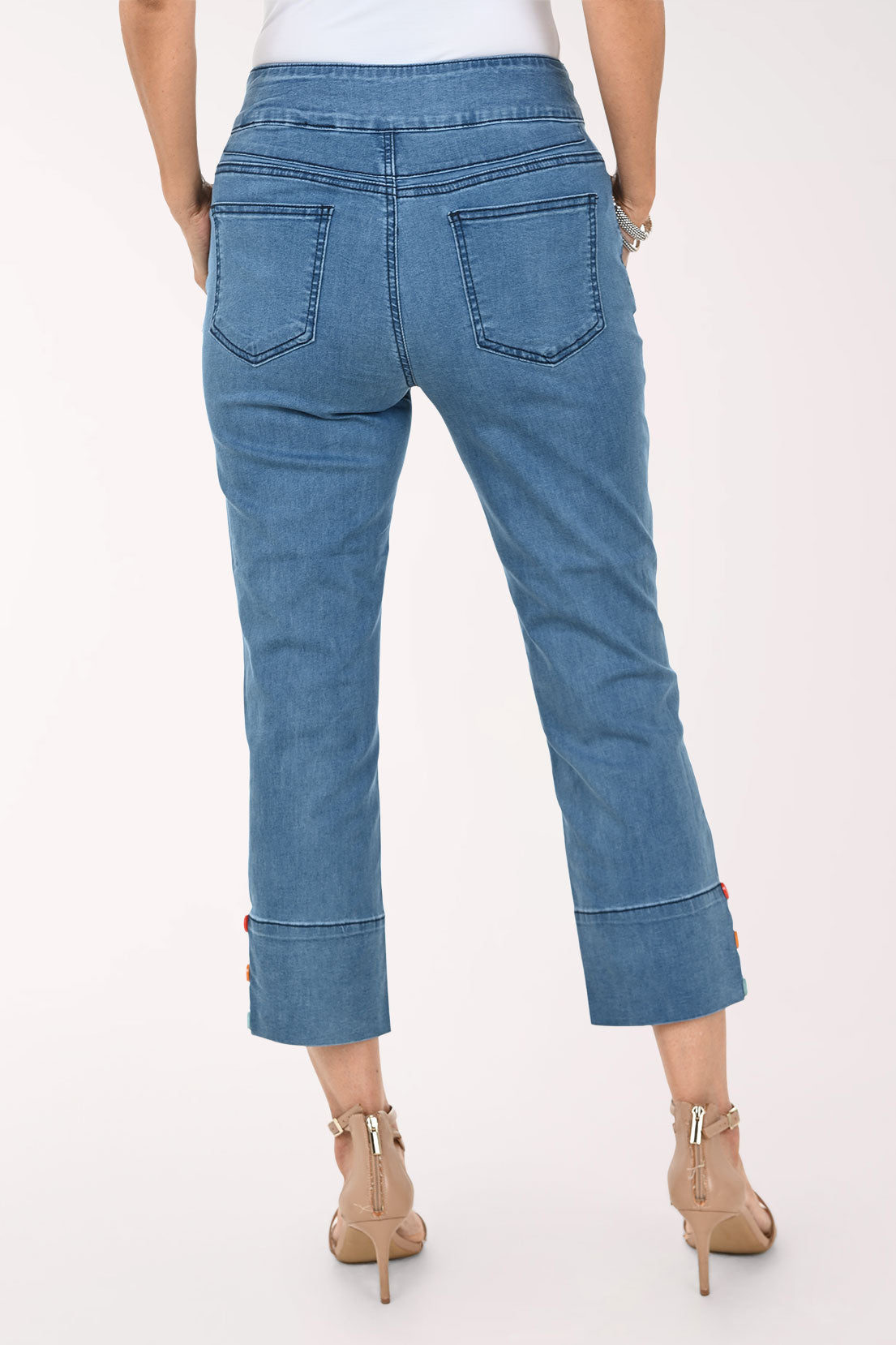 A model wearing blue cropped denim jeans with rolled cuffs featuring colorful button details, paired with beige high-heeled sandals against a plain white studio background.