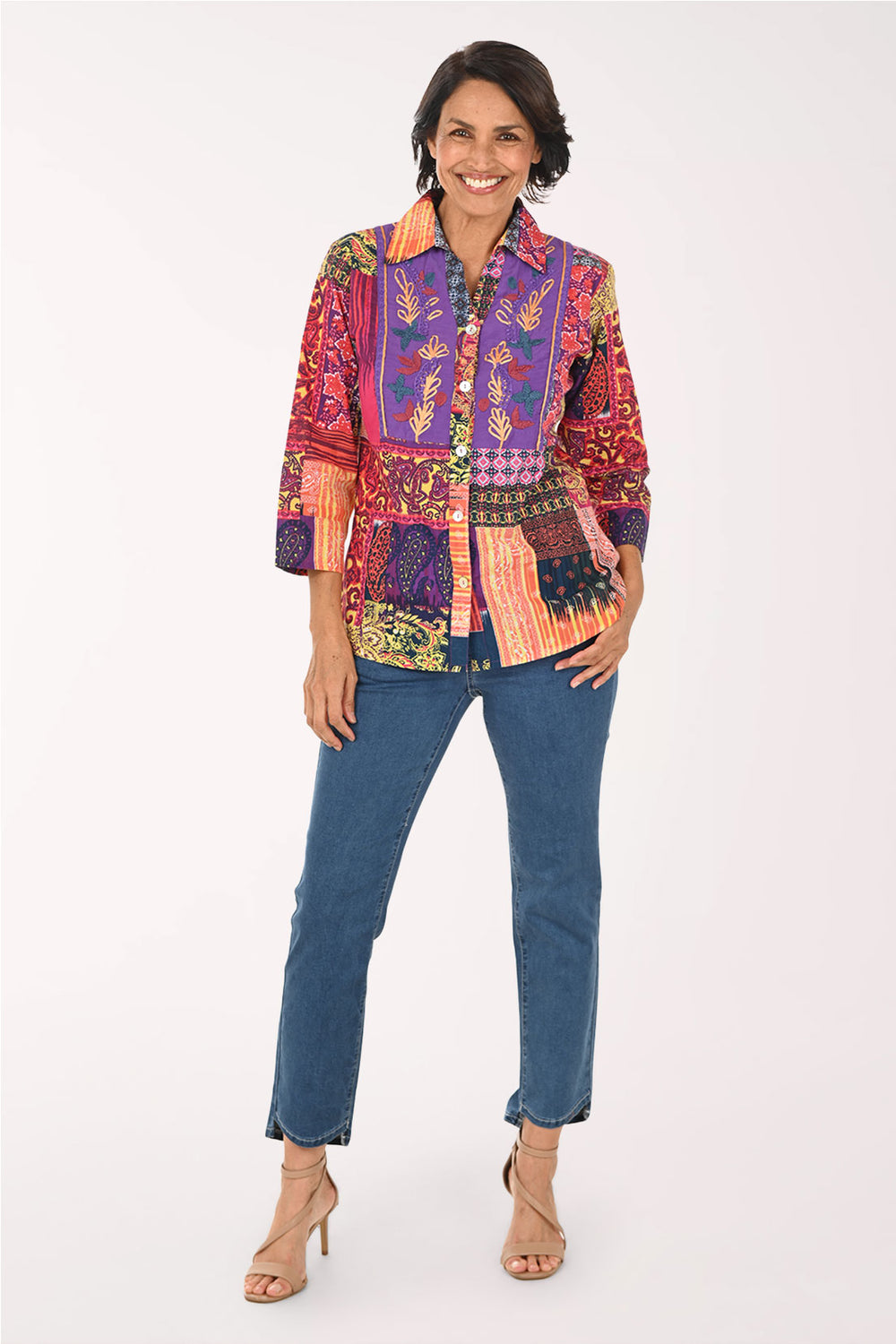 A woman modeling a vibrant, multicolored patchwork button-up shirt with blue jeans and beige high-heeled sandals, standing against a plain white studio background.