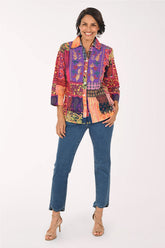 A woman modeling a vibrant, multicolored patchwork button-up shirt with blue jeans and beige high-heeled sandals, standing against a plain white studio background.