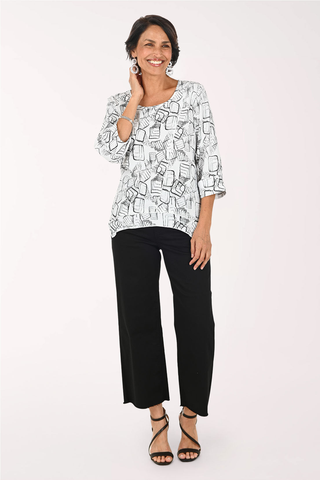 A woman modeling a three-quarter sleeve top with a black and white abstract line pattern, paired with black cropped pants and black strappy heels, against a plain white studio background.
