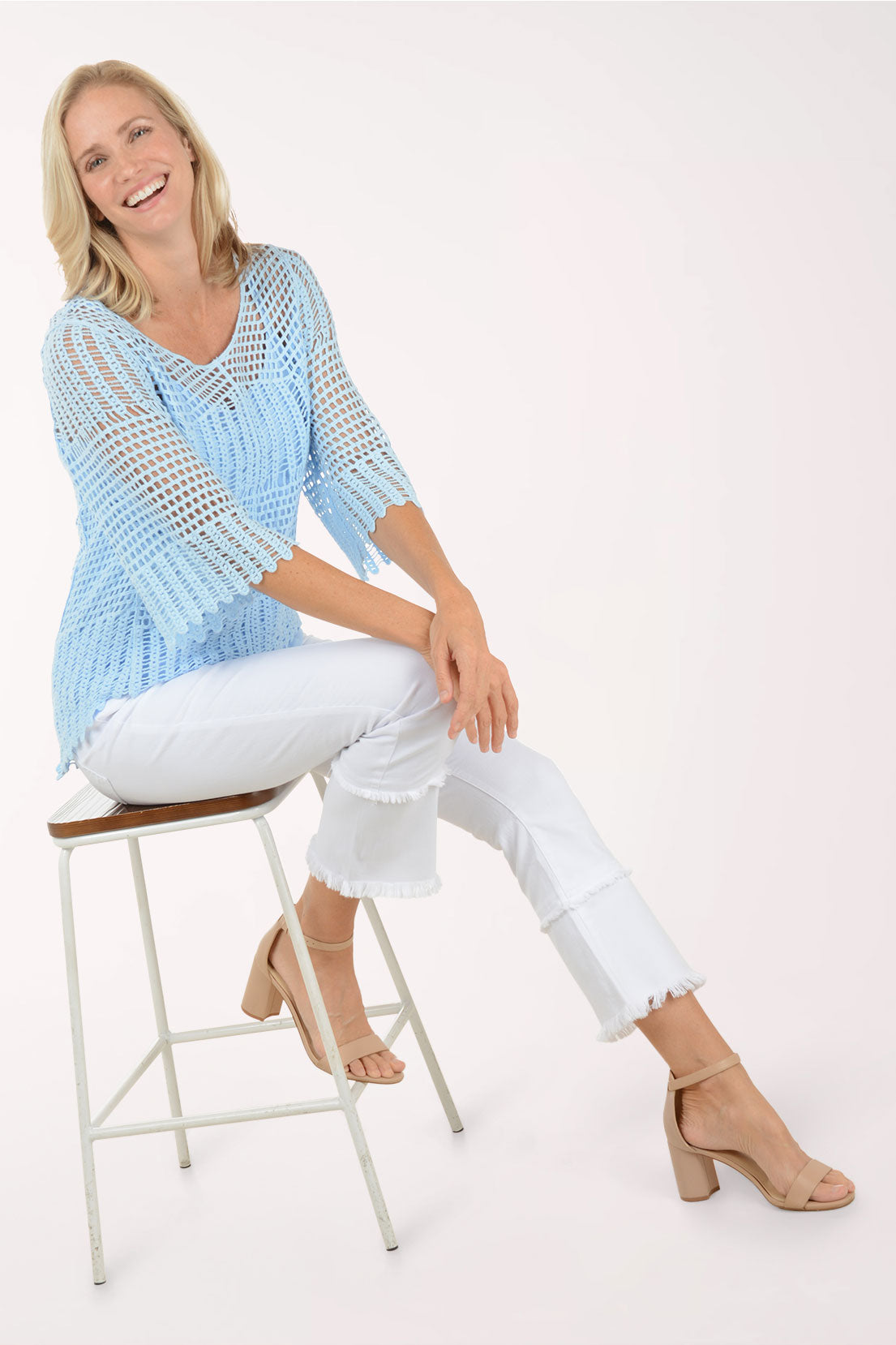 Woman wearing a light blue crochet top and white pants sitting on a stool against a white background