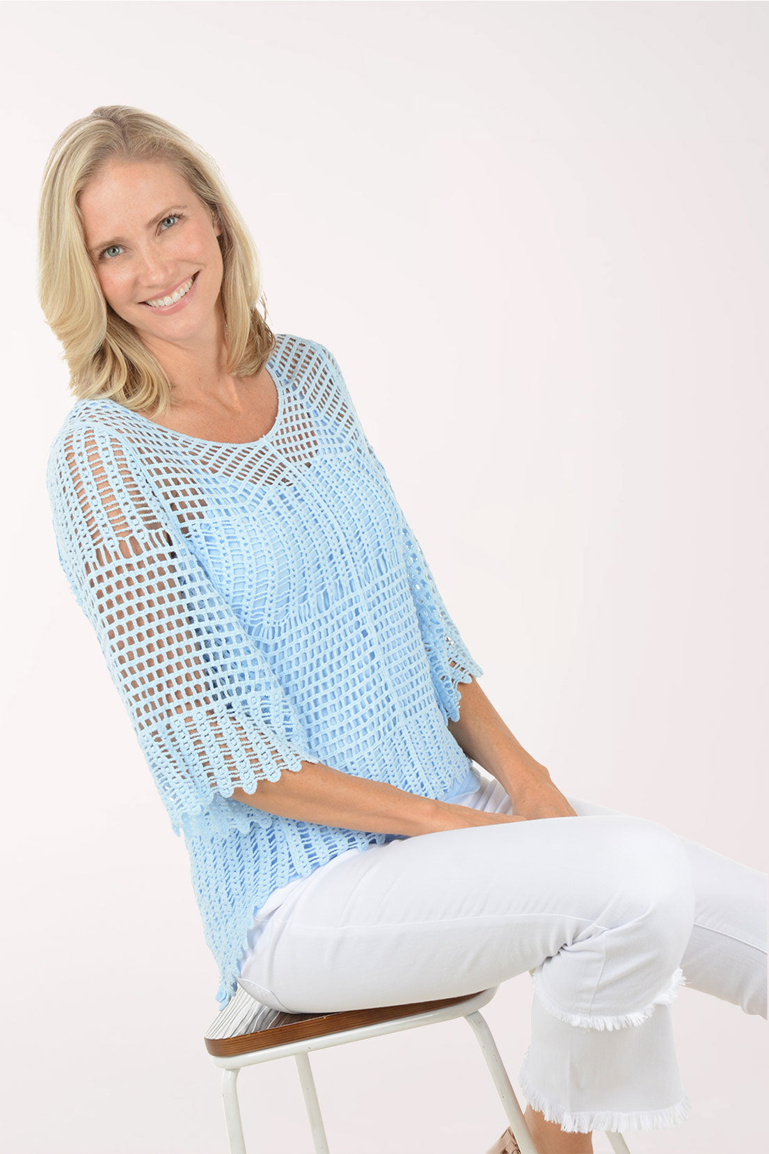 Woman wearing a light blue crochet top and white pants sitting on a stool against a white background