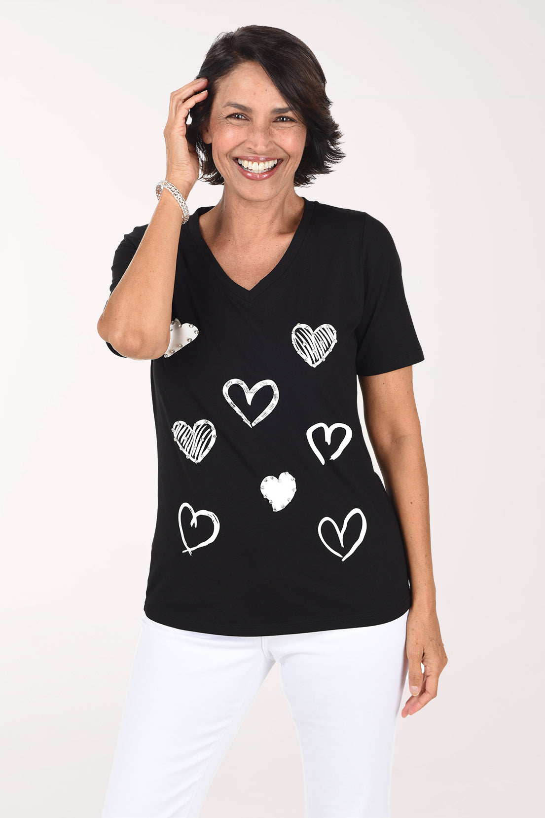 Front view of a woman in a black short sleeve V-neck tee with white and silver sketched hearts scattered across the front, some outlined and some filled, with rhinestone accents, worn with white pants.