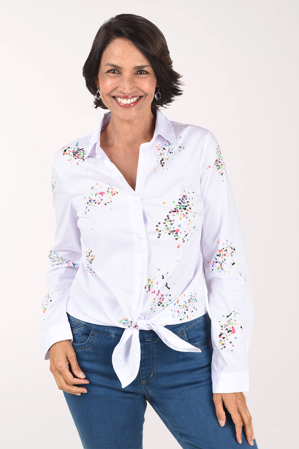 Front view of a woman in a white long sleeve button-front shirt with a classic collar and scattered clusters of multicolor paint speckles and sequins, tied at the hem over medium-wash jeans.