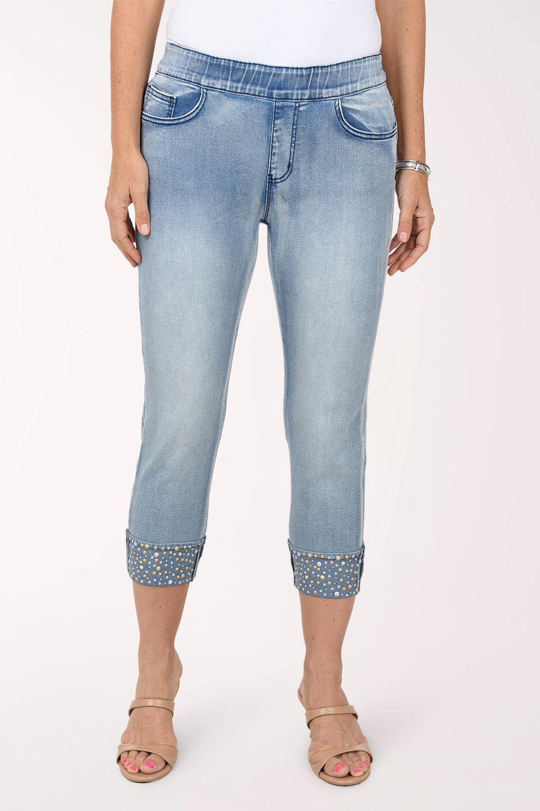 Front view of light-wash stretch denim pull-on capri pants with an elastic waistband, faux fly and cuffs studded with rhinestones and metallic studs, worn with a white top and nude sandals.