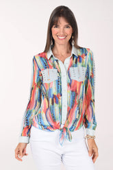 Front view of a woman in a long sleeve chiffon shirt with a coral, orange, blue and gold watercolor brushstroke print, light denim collar, placket and faux chest pockets, tied at the hem over white pants.