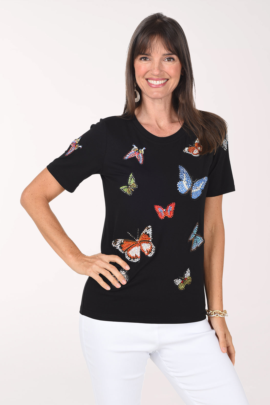 Front view of a woman in a black crew neck tee covered in rhinestone-outlined butterfly appliques in orange, blue, red, green and silver, worn with white pants.