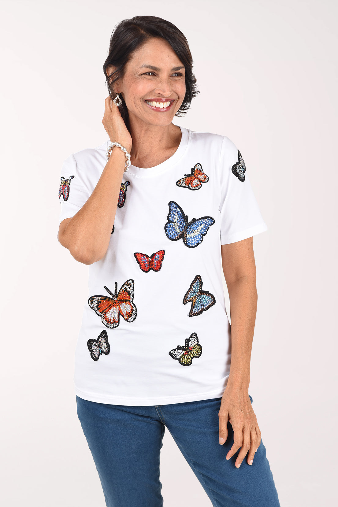 Front view of a woman in a white crew neck tee with rhinestone-outlined butterfly appliques in orange, blue, red and green, worn with medium-wash jeans.