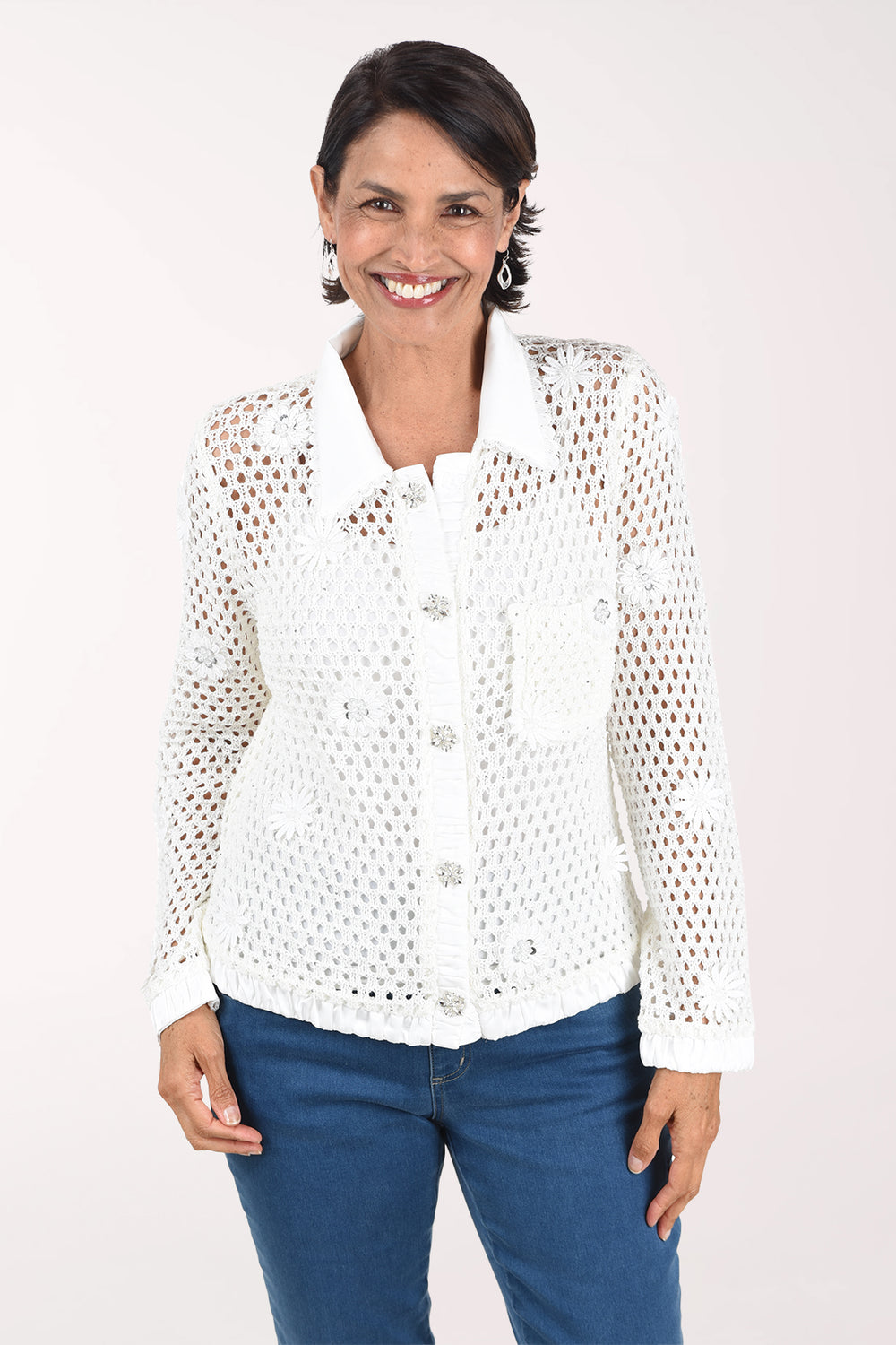 Front view of a woman in an ivory open crochet long sleeve jacket with a white satin collar, placket, cuffs and hem, jeweled floral buttons, a chest pocket and floral appliques, worn over a white cami with jeans.