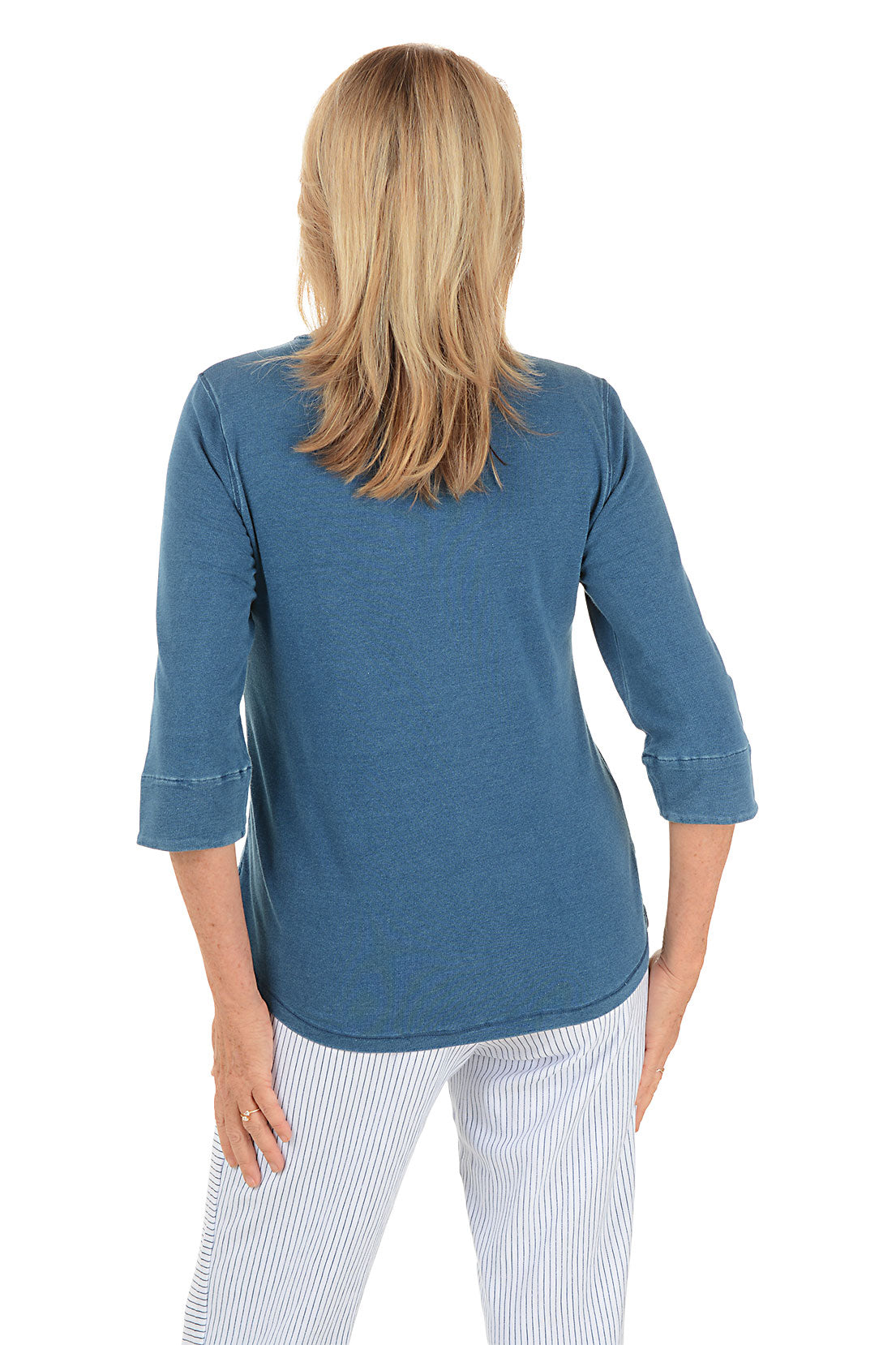 Woman wearing a blue long-sleeve shirt and white pants on a white background. Back shot.