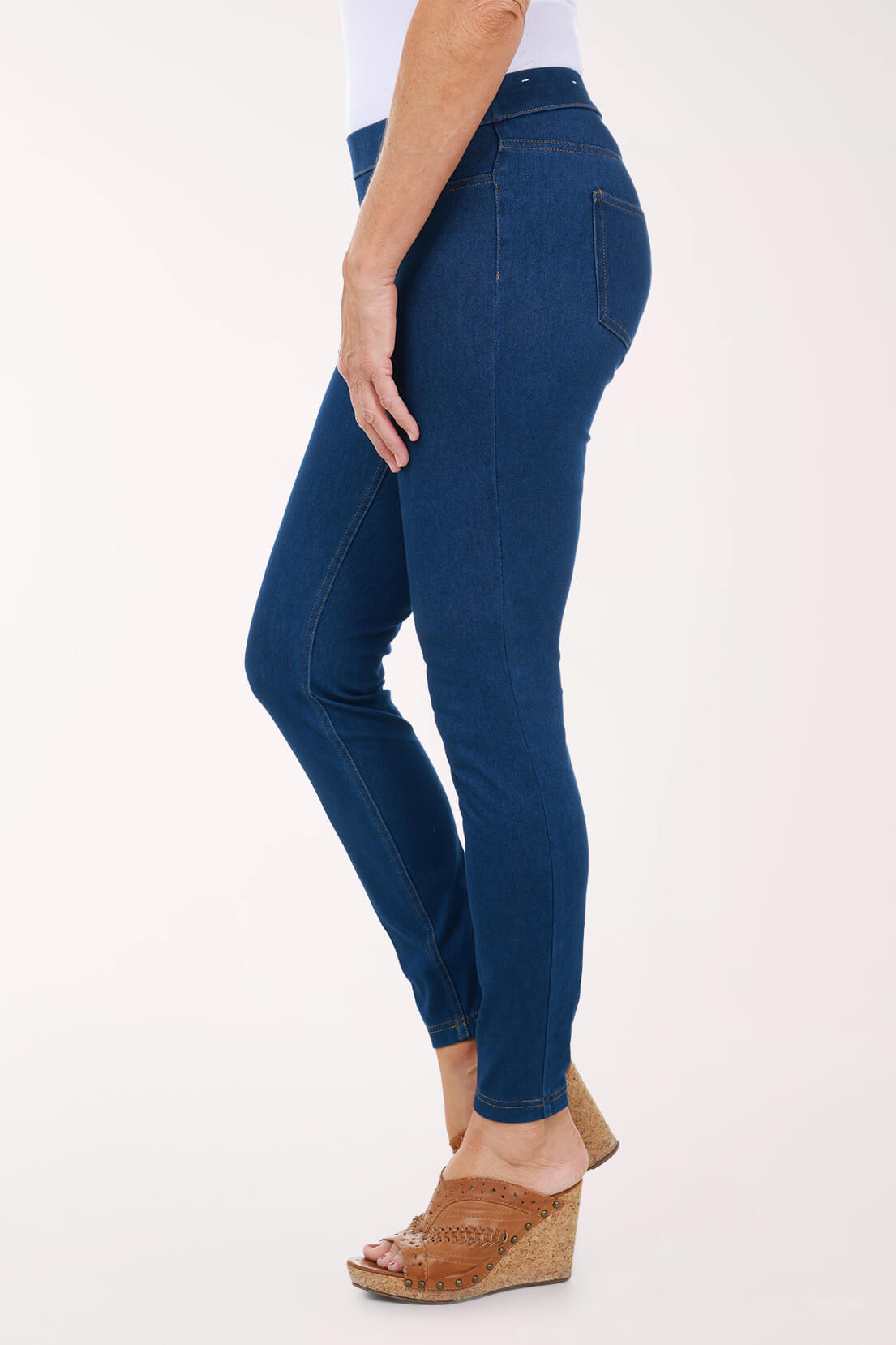 A side view of medium wash skinny pull-on jeggings showing the slim ankle-length leg, worn with tan wedge sandals.