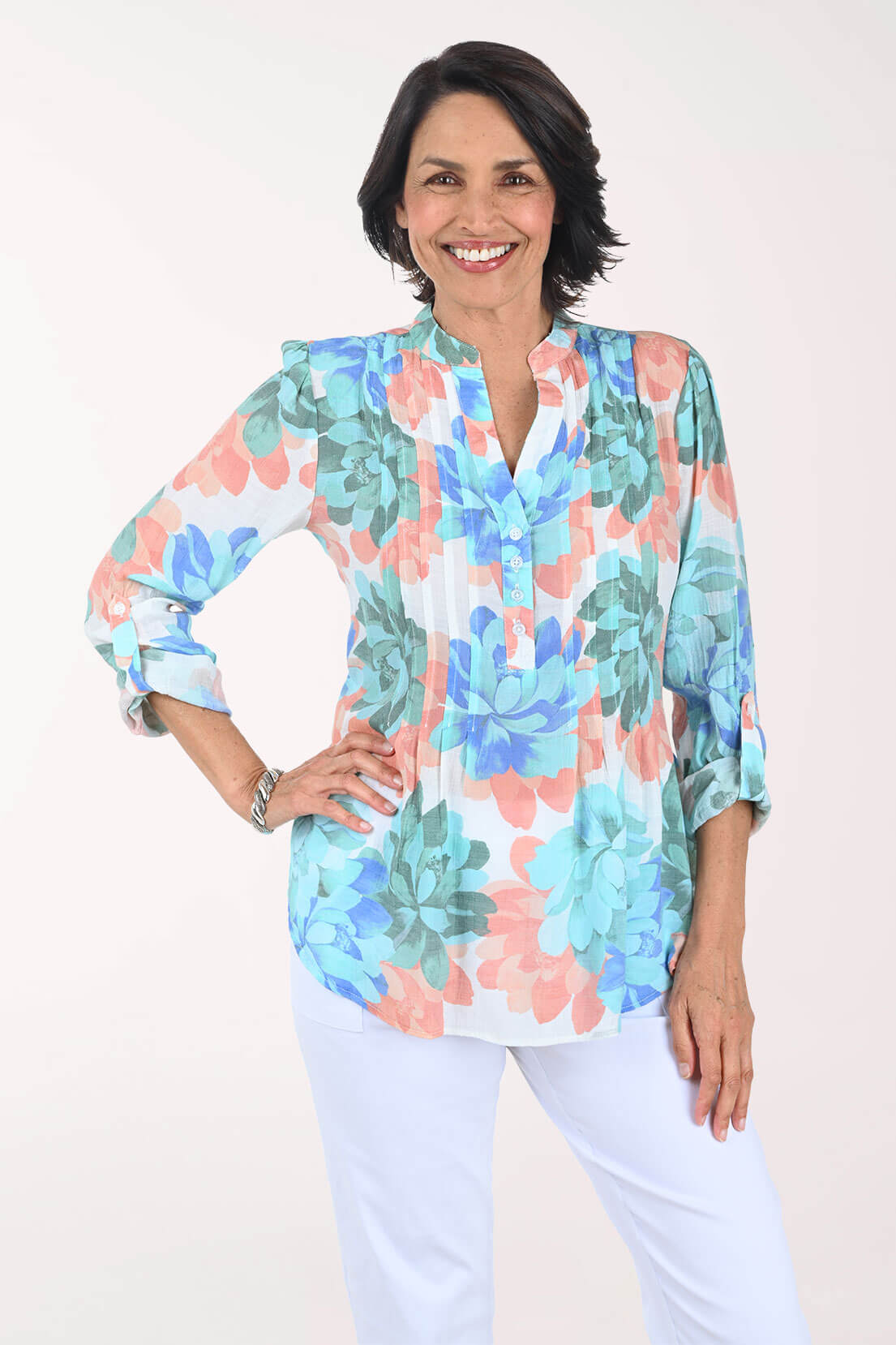 Woman smiling with one hand on her hip in a white blouse printed with large watercolor flowers in turquoise, cobalt blue, coral, and sage green, with a mandarin collar, split V-neck, and sleeves rolled to the elbow, worn with white pants.