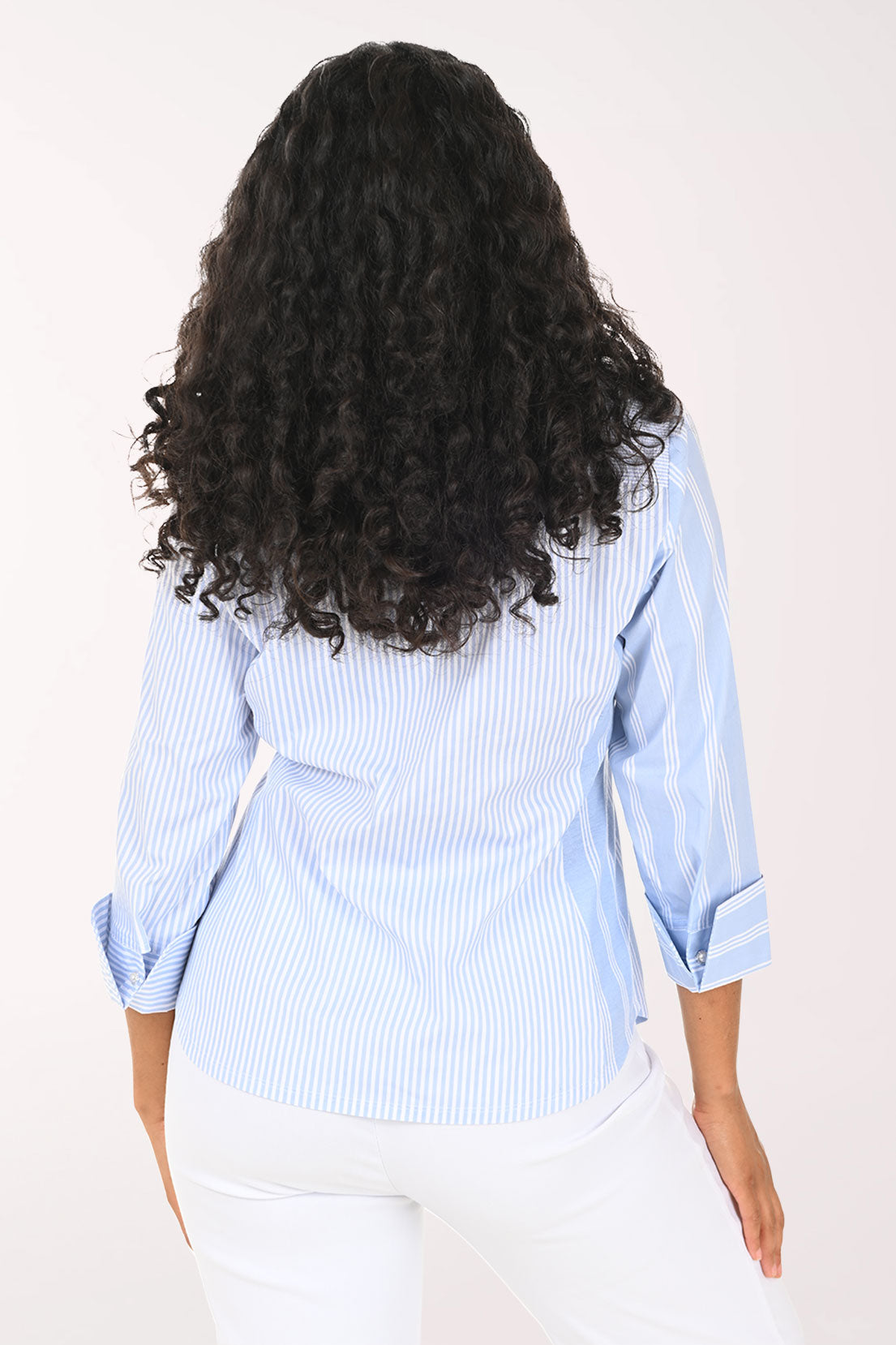A smiling woman with long curly black hair wearing a light blue and white vertically striped button-down shirt with white pants, standing against a plain white studio background.