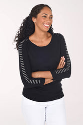 A smiling woman with curly dark hair wearing a black long-sleeve top with white dotted trim along the sleeves and white pants, standing against a plain light background.
