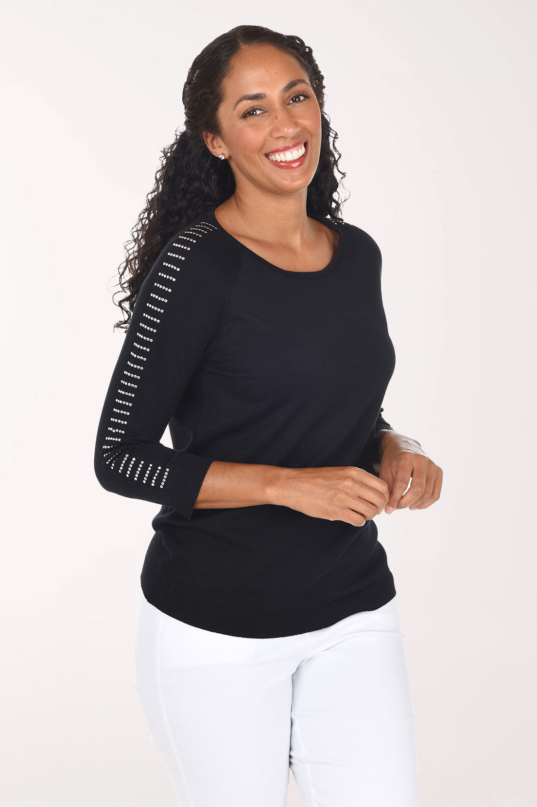 A smiling woman with curly dark hair wearing a black long-sleeve top with white dotted trim along the sleeves and white pants, standing against a plain light background.
