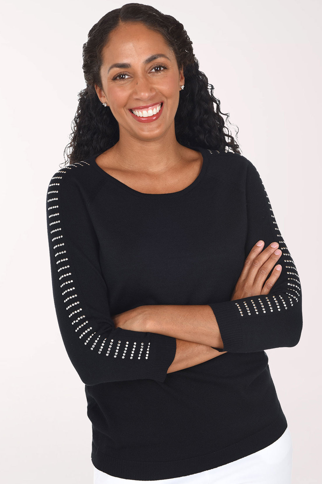 A smiling woman with curly dark hair wearing a black long-sleeve top with white dotted trim along the sleeves and white pants, standing against a plain light background.