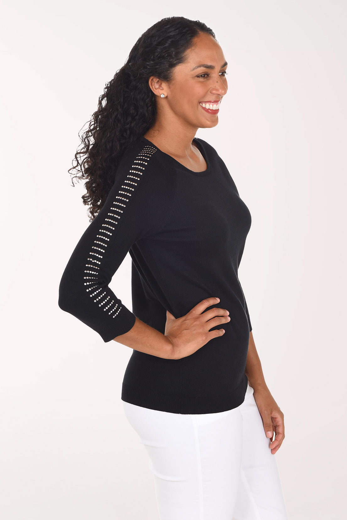 A smiling woman with curly dark hair wearing a black long-sleeve top with white dotted trim along the sleeves and white pants, standing against a plain light background.