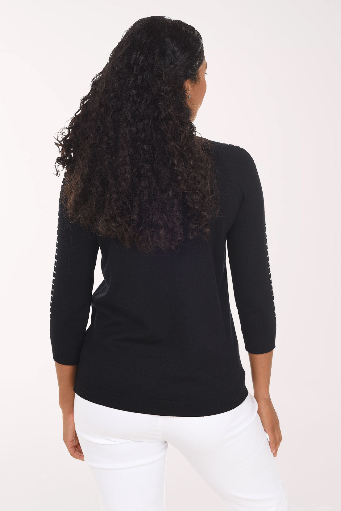 A smiling woman with curly dark hair wearing a black long-sleeve top with white dotted trim along the sleeves and white pants, standing against a plain light background.