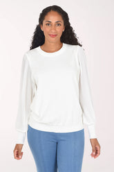 A woman modeling a white long-sleeve crewneck sweater with textured, pleated sheer sleeves, paired with blue jeans, against a plain light background.