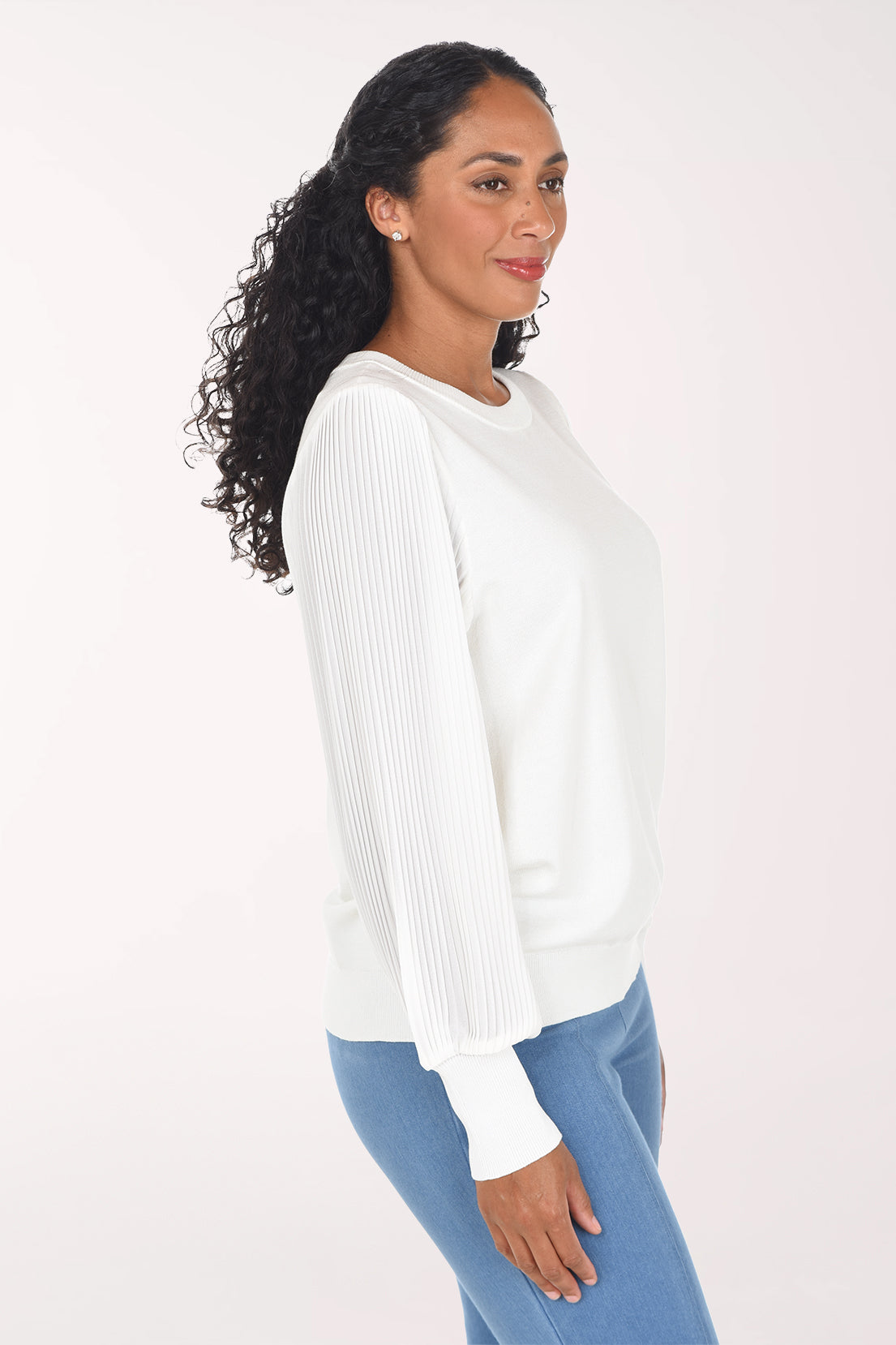 A woman modeling a white long-sleeve crewneck sweater with textured, pleated sheer sleeves, paired with blue jeans, against a plain light background.