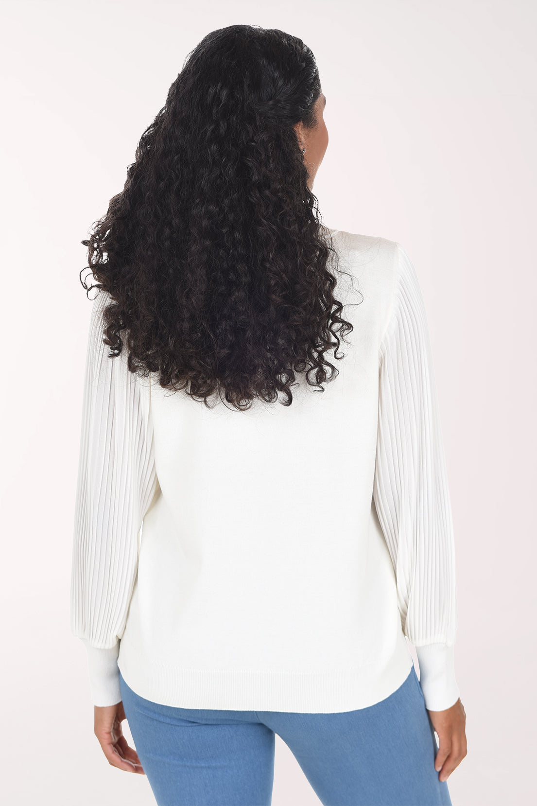 A woman modeling a white long-sleeve crewneck sweater with textured, pleated sheer sleeves, paired with blue jeans, against a plain light background.