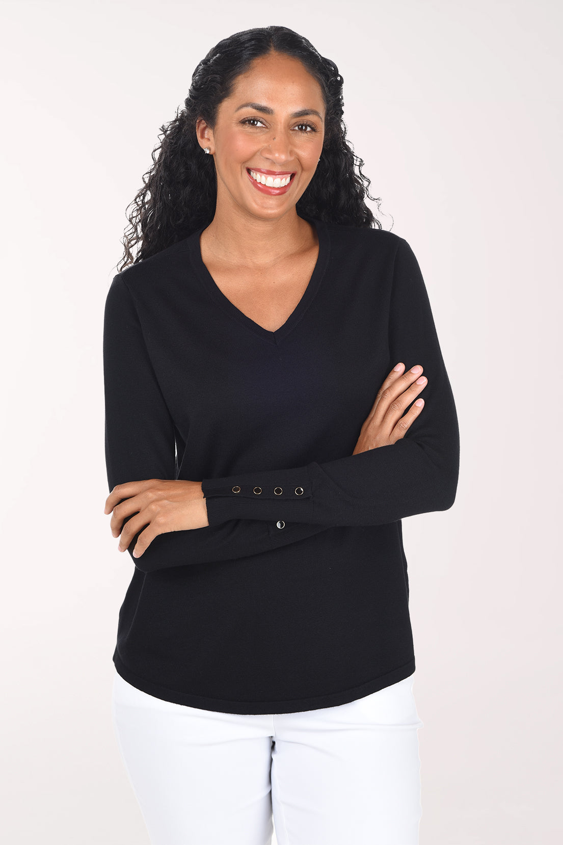 A smiling woman with long curly black hair wearing a black long-sleeve V-neck top with decorative buttons on the cuff and white pants, standing against a plain light background.