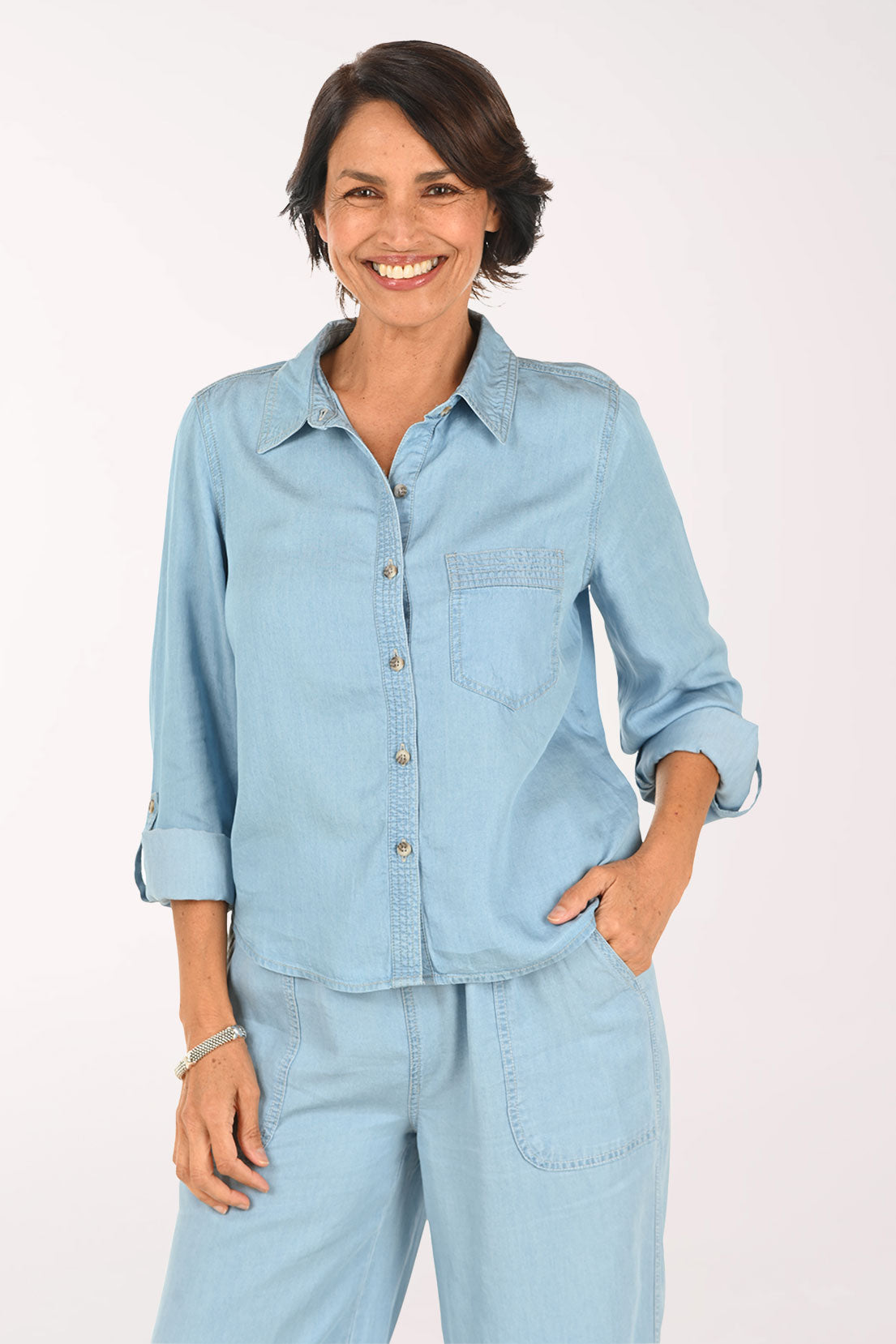 A smiling woman wearing a light blue denim button-up shirt and matching light blue denim pants, photographed against a plain light background.